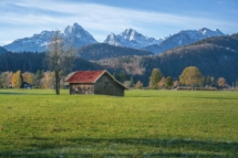 Holzhaus mit rotem Dach auf einem Feld mit den Tannheimer Alpen - Schwangau, Bayern, Deutschland